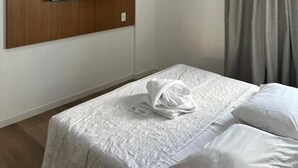 Habitación de hotel con televisor de pantalla plana montado en un panel de madera, cama con ropa blanca y unidad de aire acondicionado en la pared.