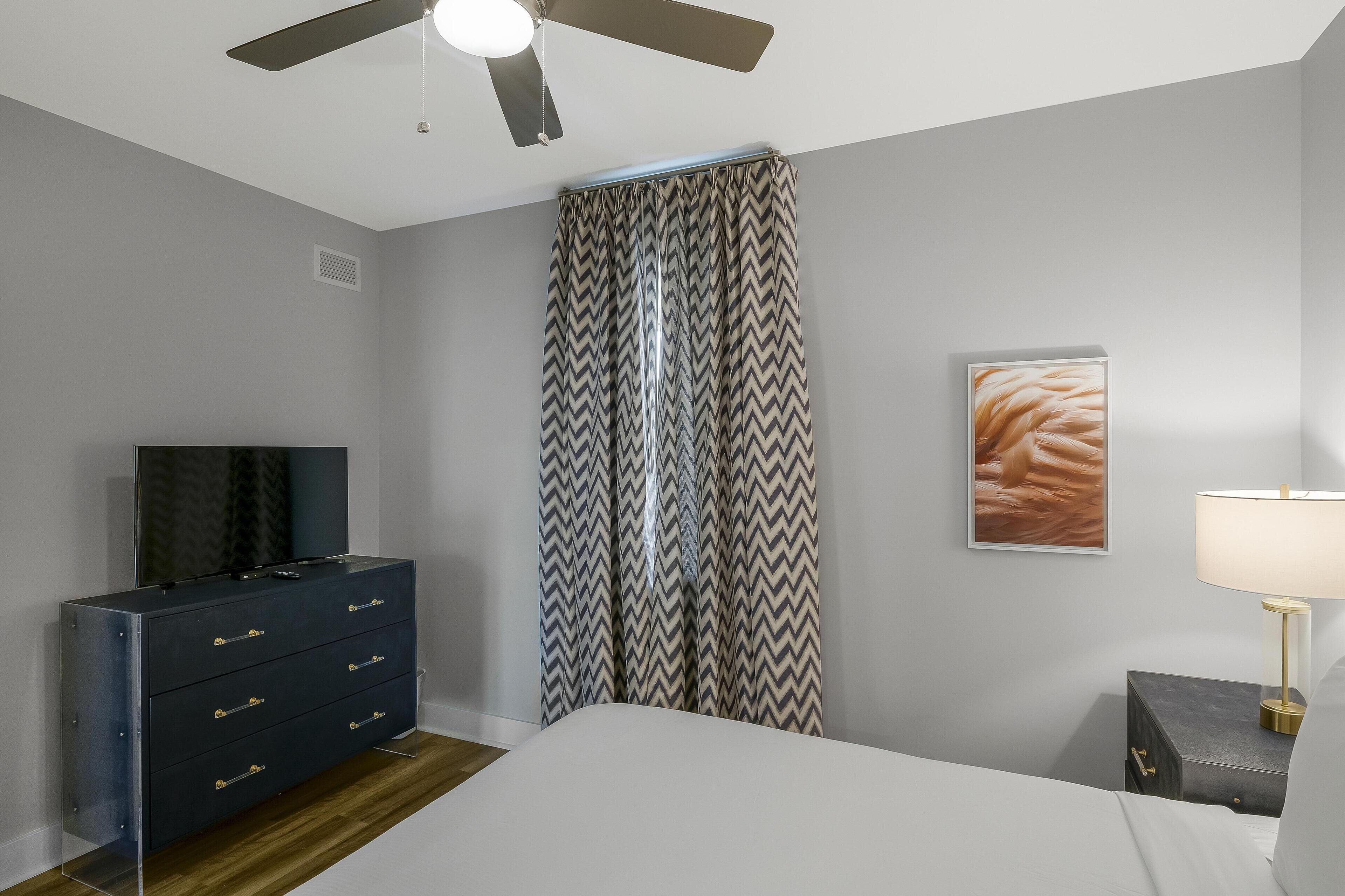 A bedroom with a bed, a dresser, a ceiling fan, a window with curtains, and a bedside lamp.