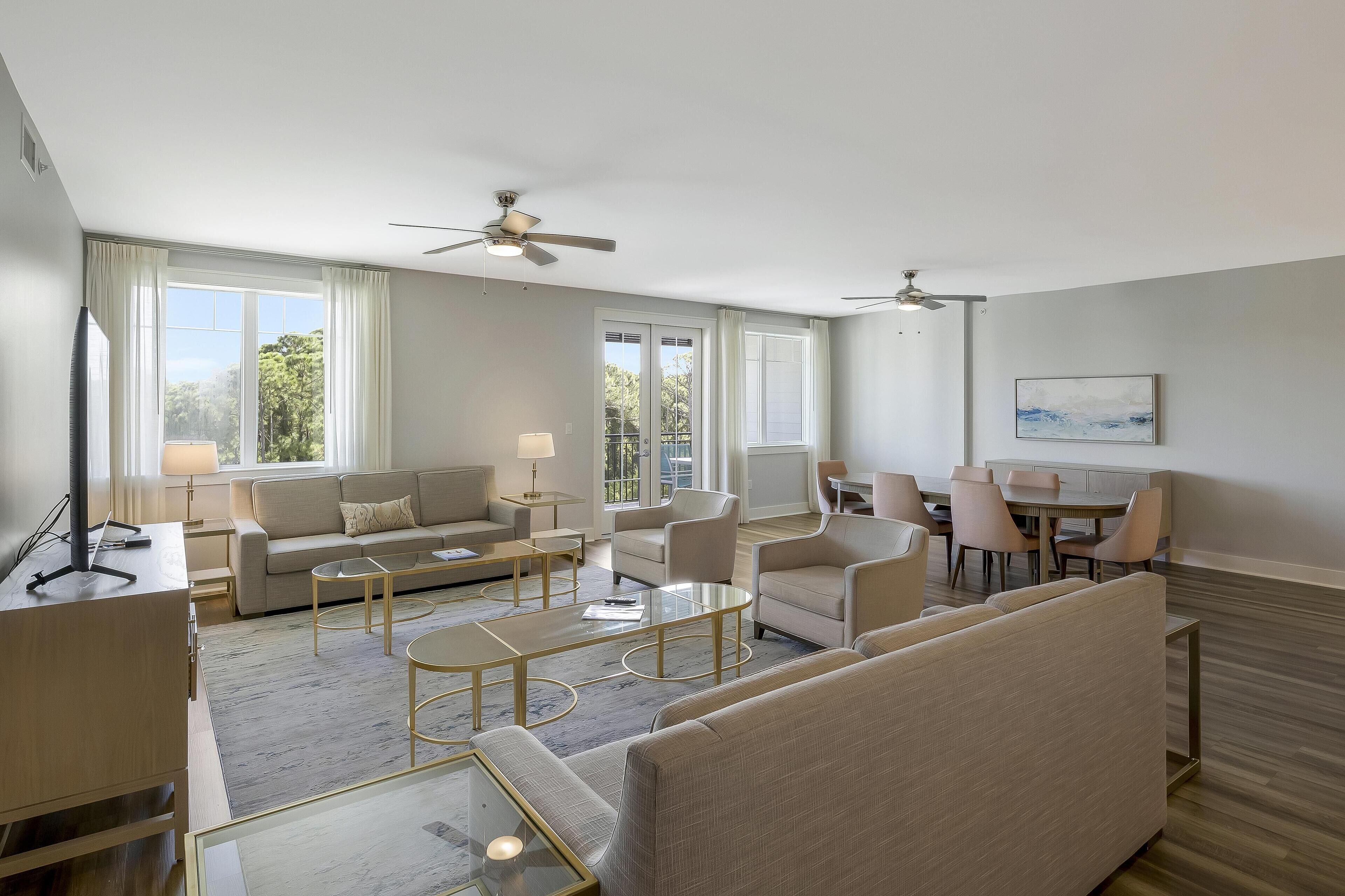 A spacious living room with a sofa, coffee table, and chairs, a dining area with a table and chairs, and large windows with curtains.