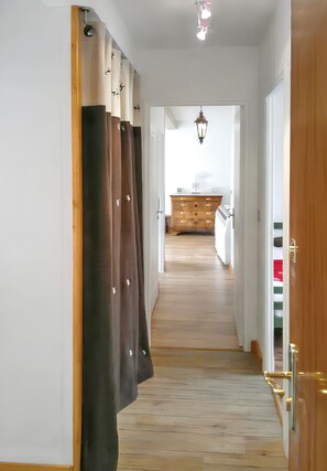 Un corridoio con pavimento in legno, una porta in legno sulla destra e vista su una stanza con una cassettiera in legno e una lampada a sospensione.