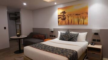 A hotel room with a bed, a desk, a chair, a small table, and a painting on the wall.