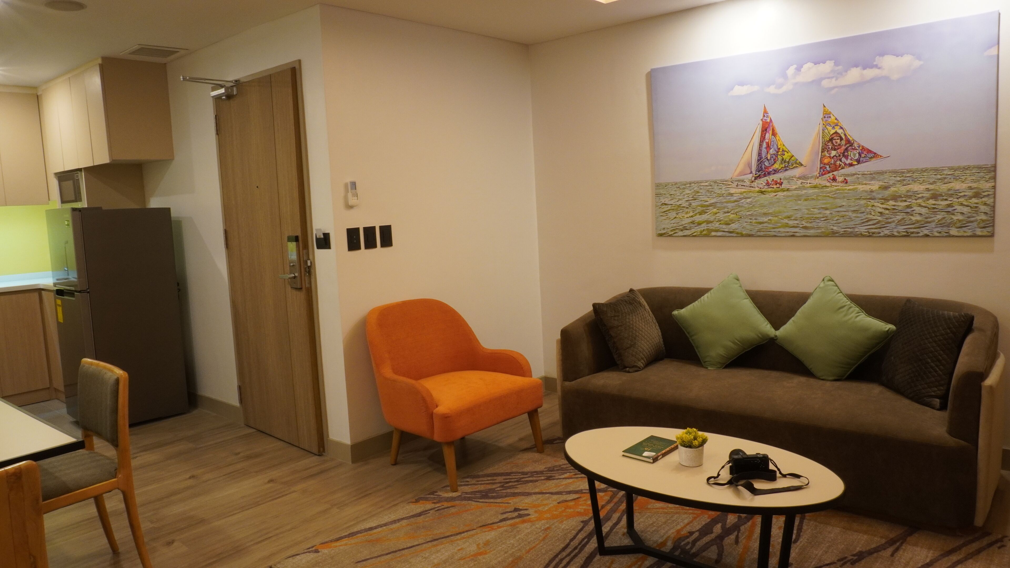 Ein gemütlicher Wohnraum mit Küchenzeile, einer Couch mit Kissen, einem kleinen Tisch mit Pflanze und Kamera sowie einem Segelbootgemälde an der Wand.