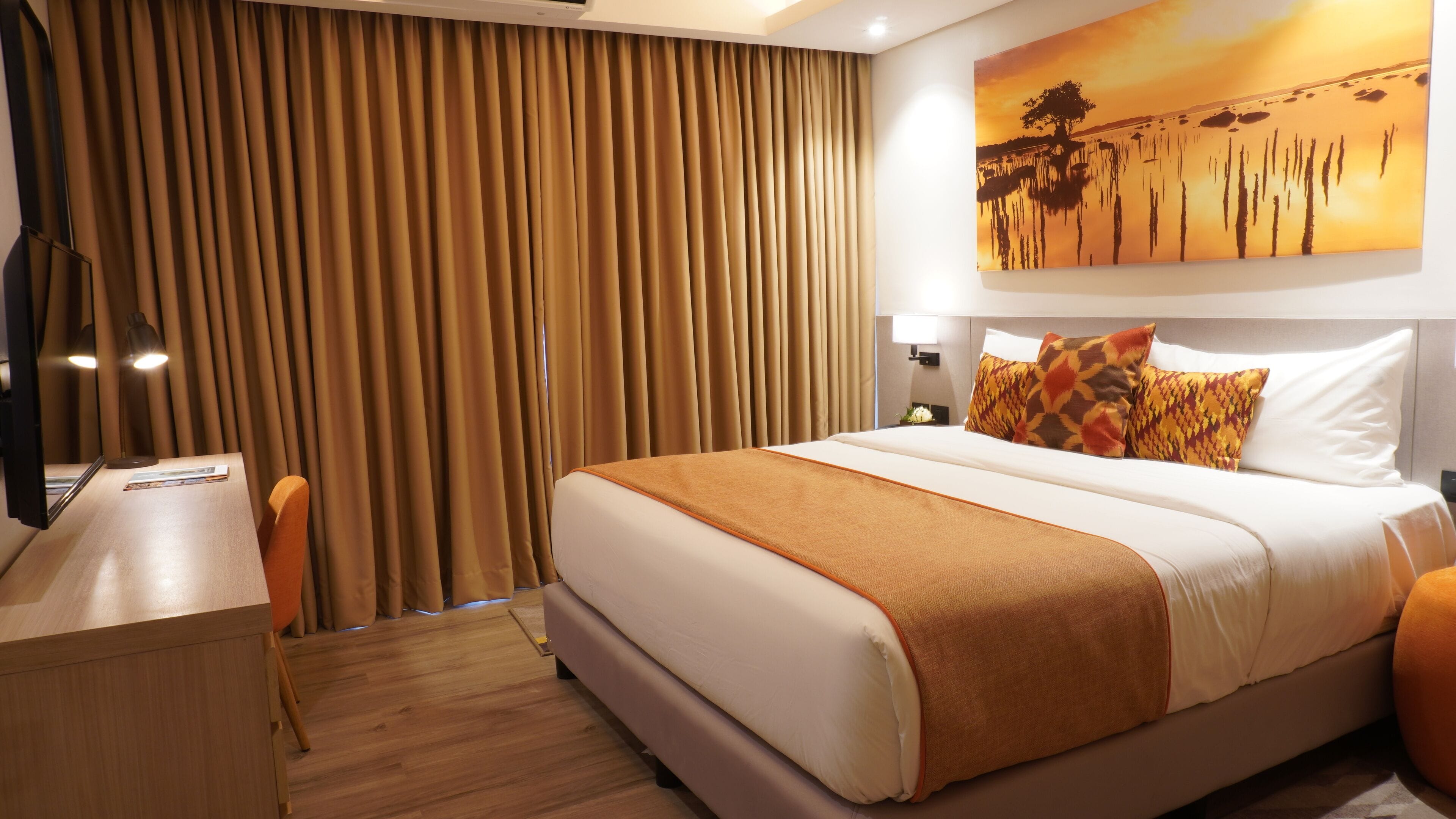 A hotel room with a large bed, a desk with a chair, a nightstand, and a painting on the wall.
