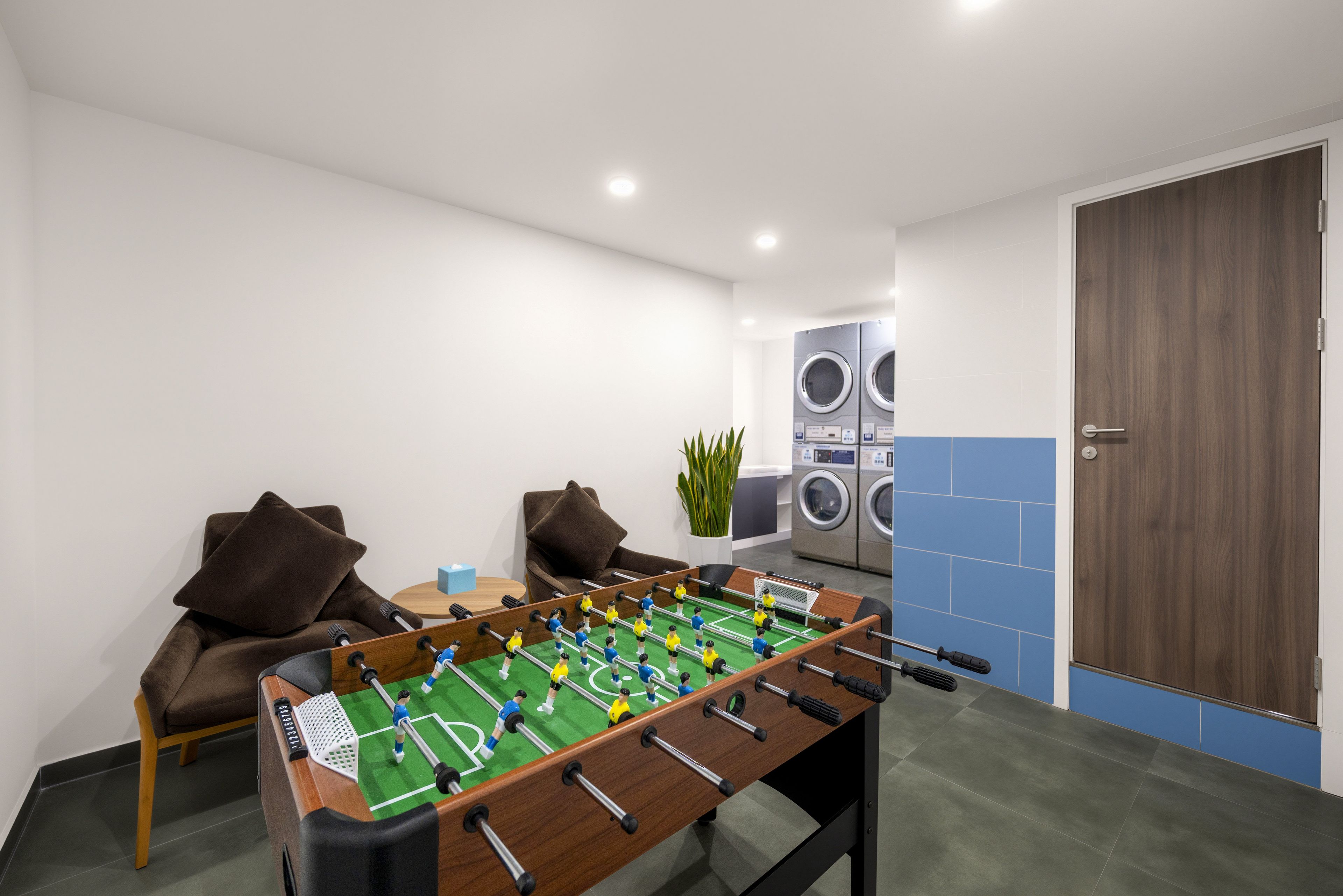 Una habitación con mesa de futbolín, sofá, una mesita con un objeto azul y una lavadora al fondo.