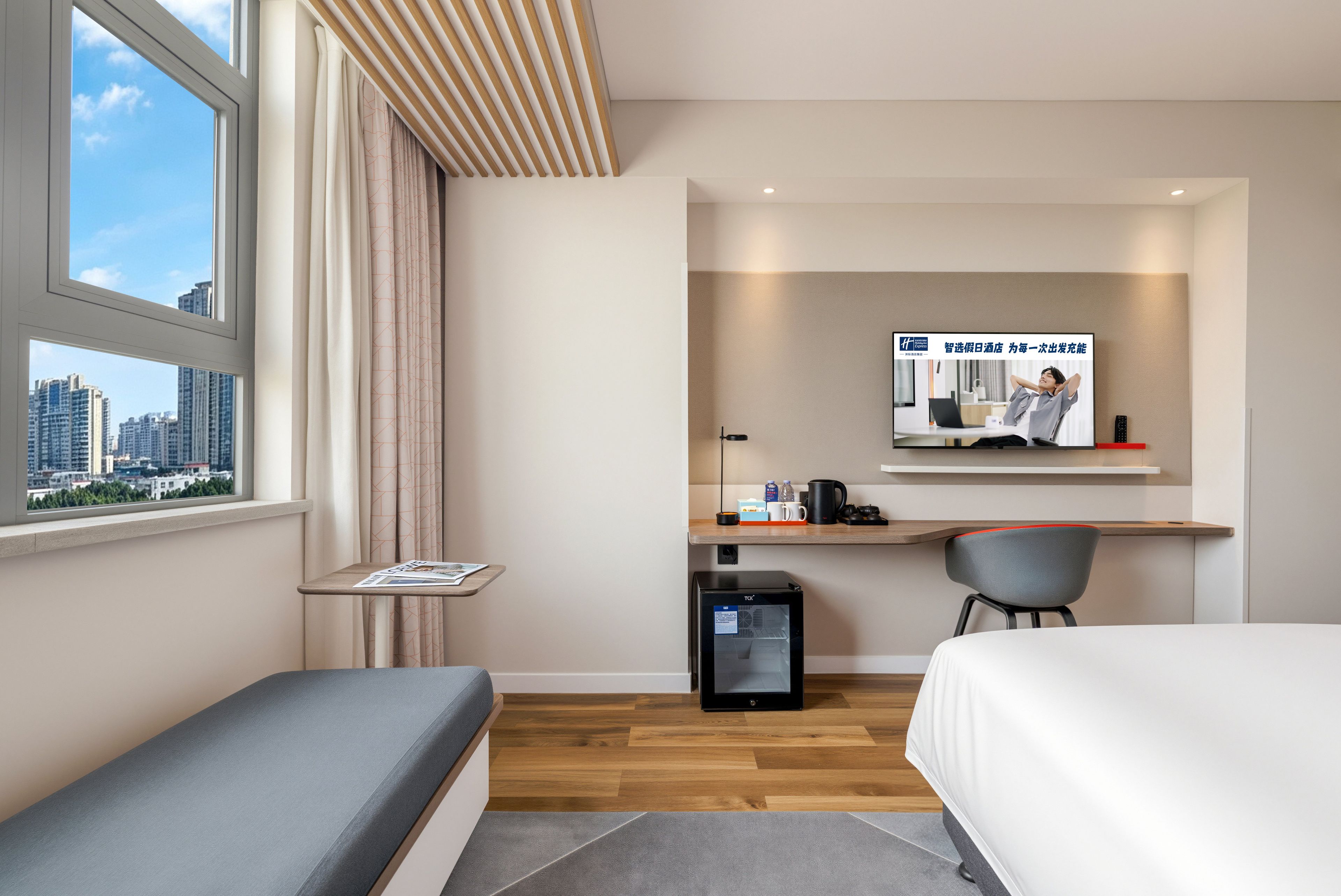 A modern hotel room with a bed, a desk, a chair, a small fridge, and a TV displaying a man relaxing.