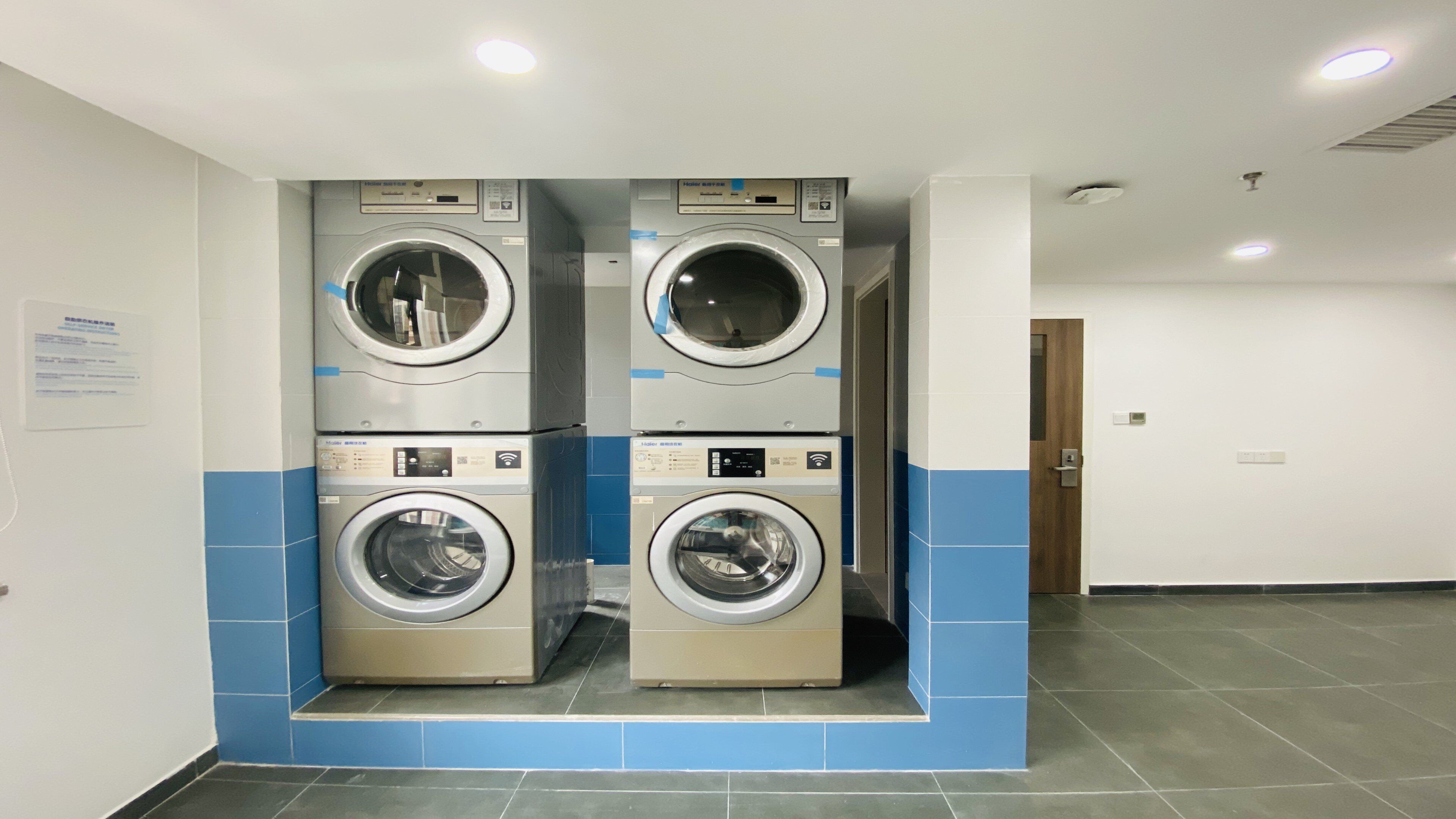 A laundry facility with four stacked washing machines, a blue-tiled floor, and a door in the background.