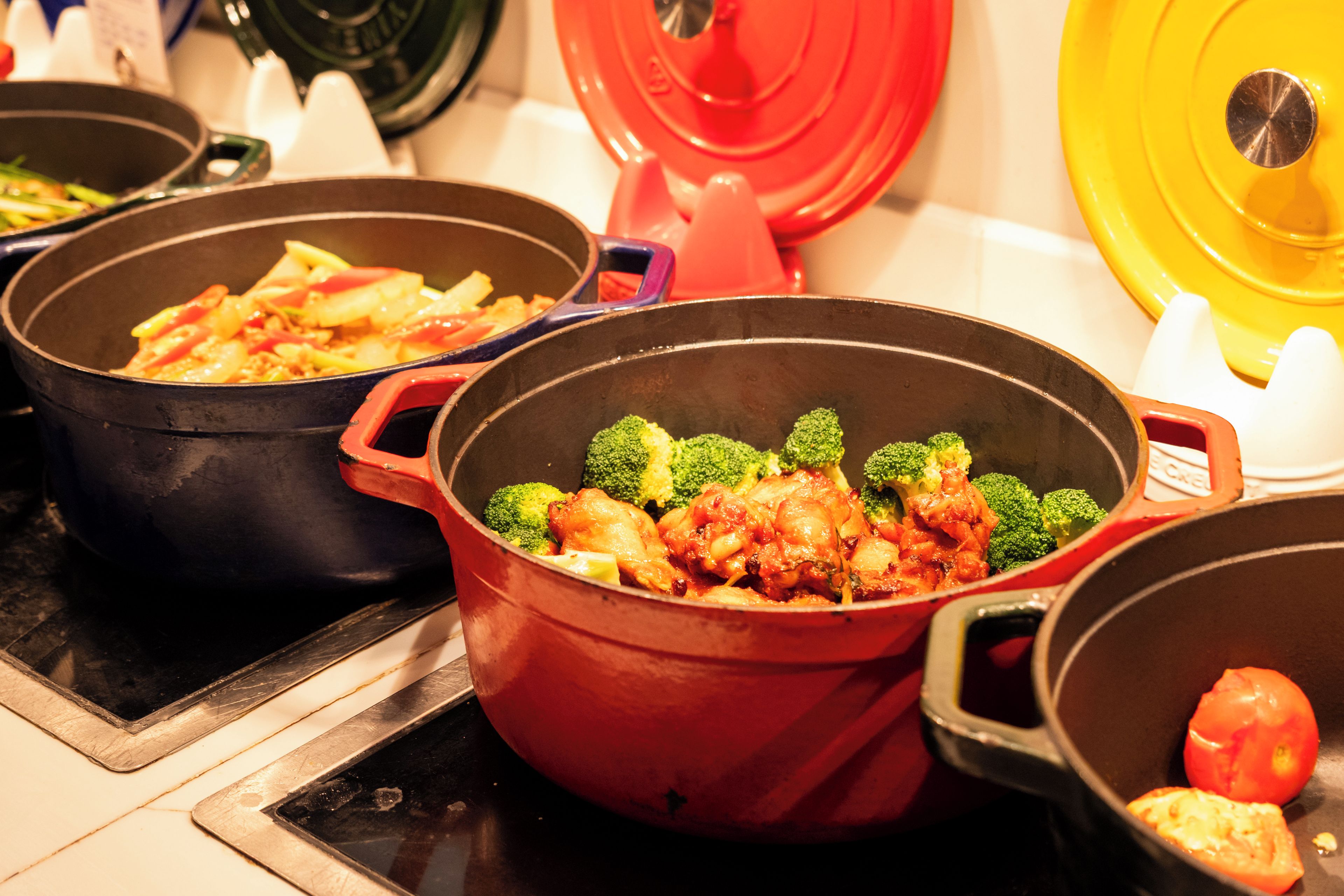 Una cocina con varias ollas que contienen comida, una olla roja con brócoli y una amarilla con tomate.