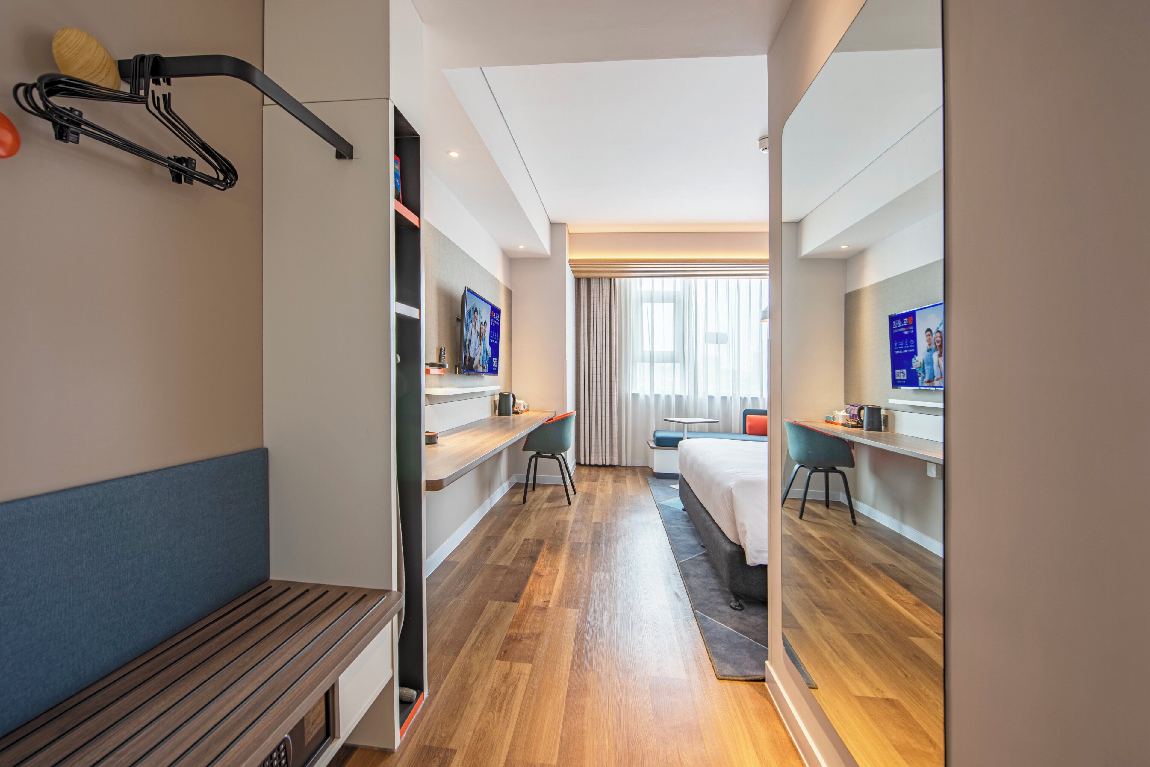 A hotel room with a bed, a desk, a chair, a bench, a TV, and a window with curtains.