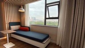 Une chambre avec un lit simple, donnant sur un paysage urbain grâce à une grande fenêtre, comprenant une table de chevet et une lampe suspendue.