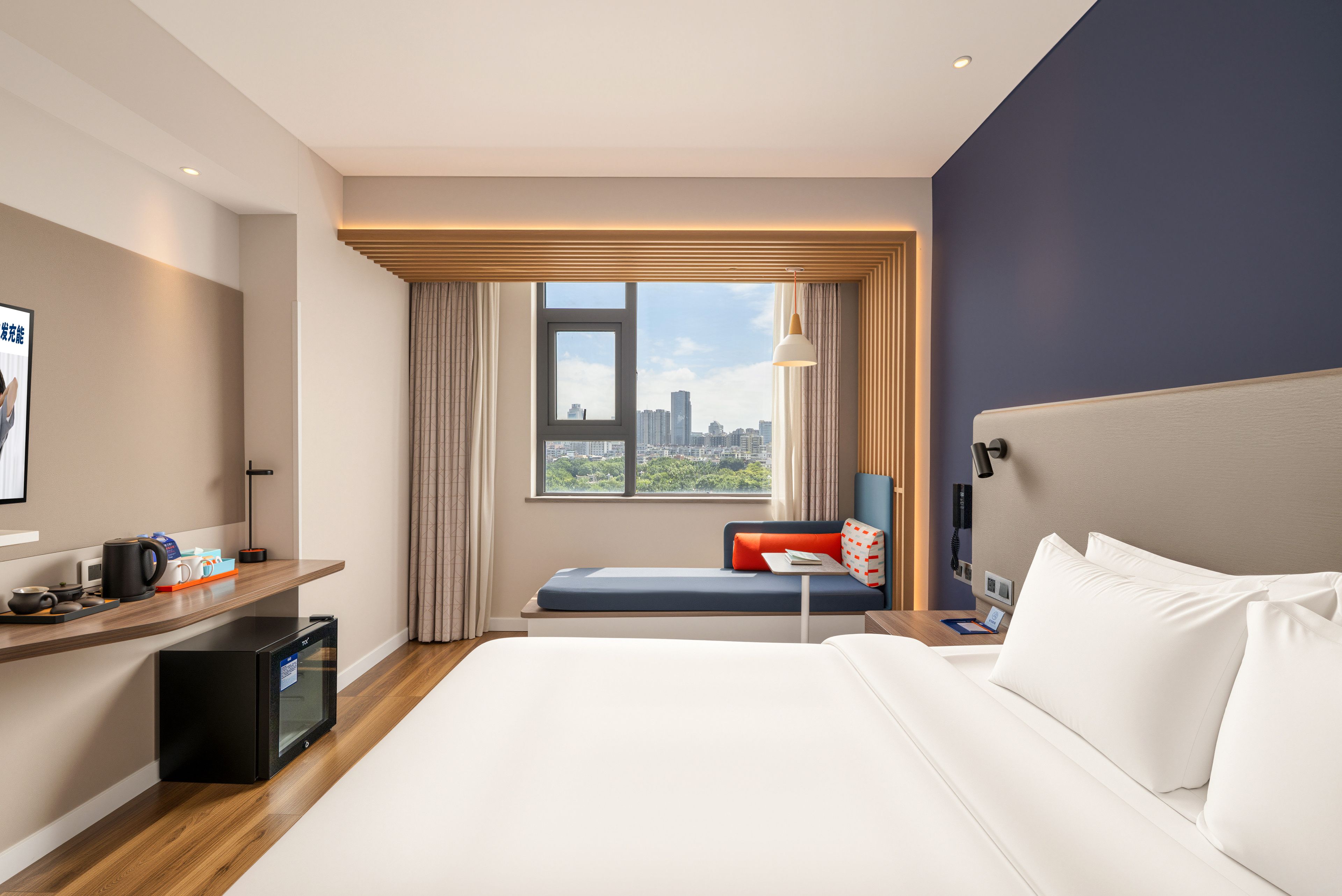 Quarto de hotel com cama, escrivaninha, cadeira, janela com vista para a cidade e pequena mesa com luminária.