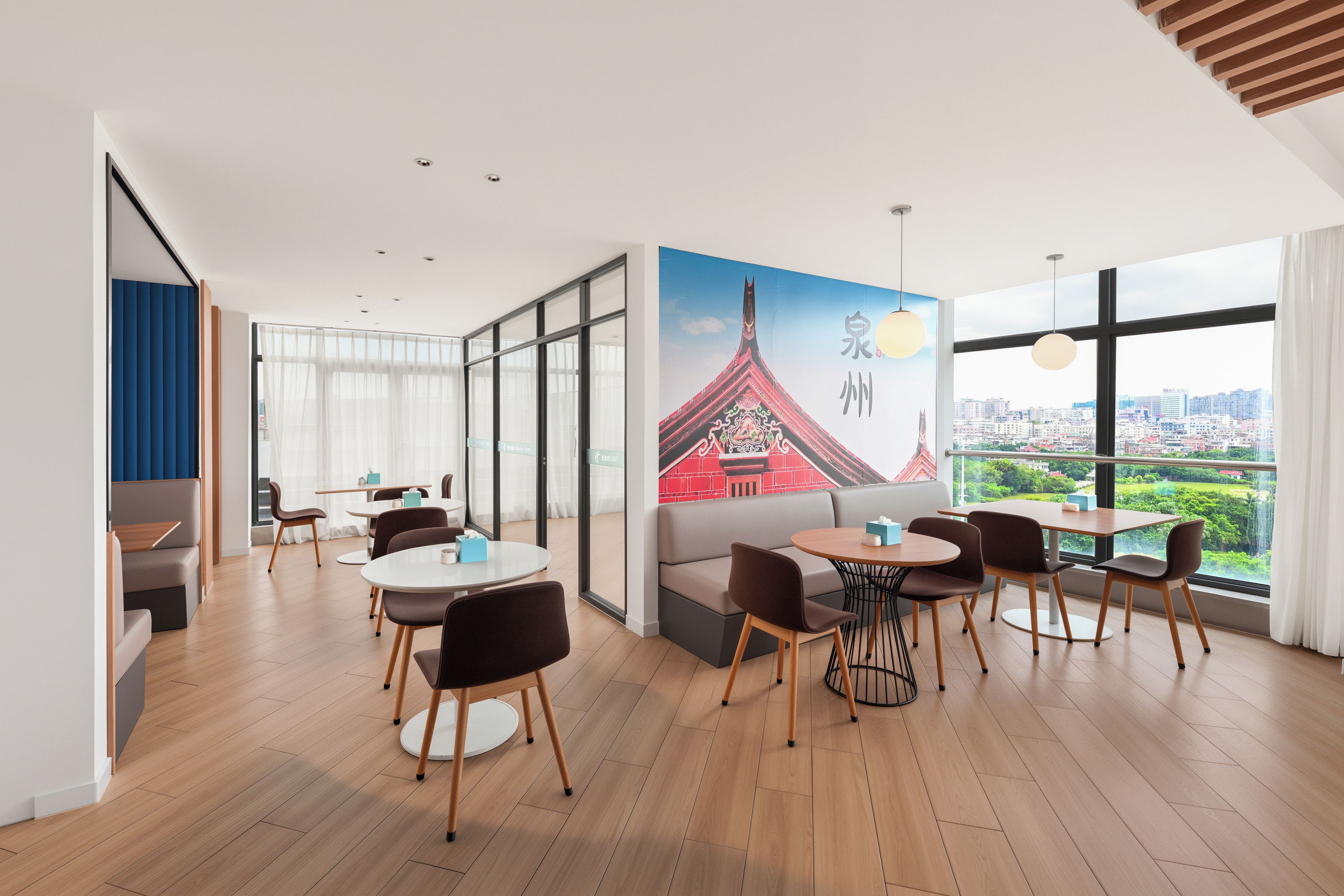 Ein modernes Hotelzimmer mit großem Fenster und Blick auf die Stadt, einer Essecke mit runden Tischen und Stühlen sowie einer Wand mit einer Wandmalerei eines traditionellen Gebäudes.