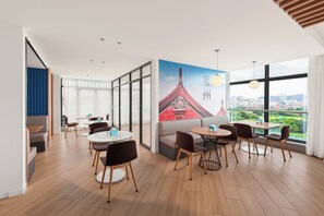 A modern hotel room with a large window offering a city view, a dining area with round tables and chairs, and a wall with a traditional building mural. - Holiday Inn Express Quanzhou City Center by IHG (Quanzhou)