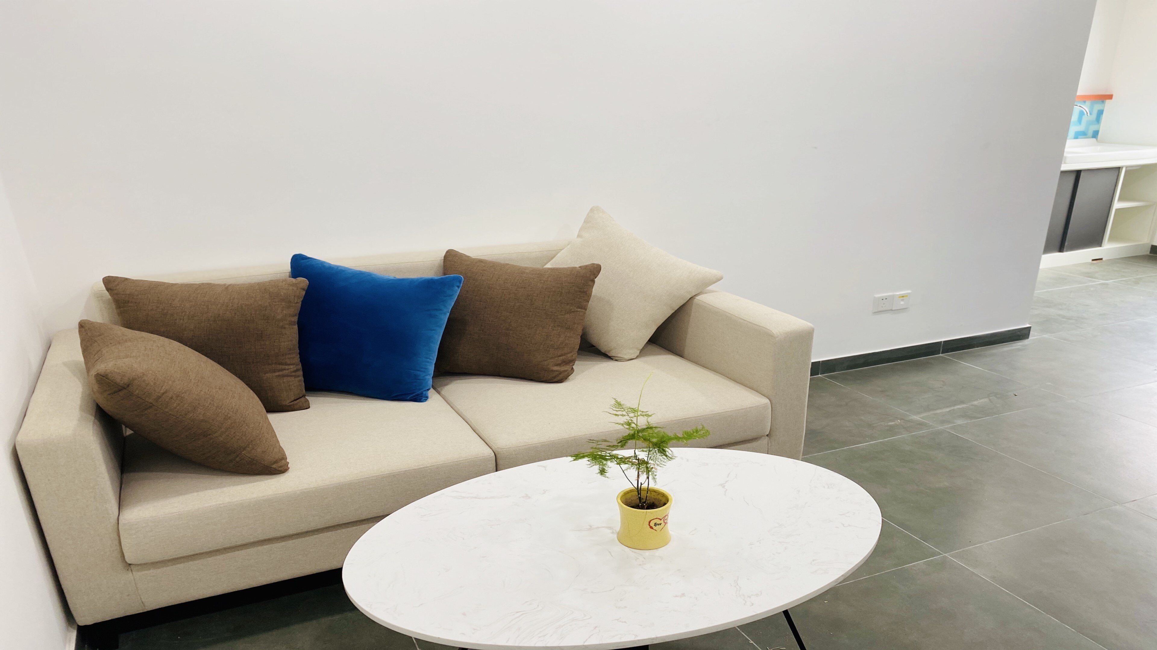 Una sala de estar moderna con un sofá color beige, cojines marrones y azules, y una mesa de centro redonda blanca con una planta pequeña.