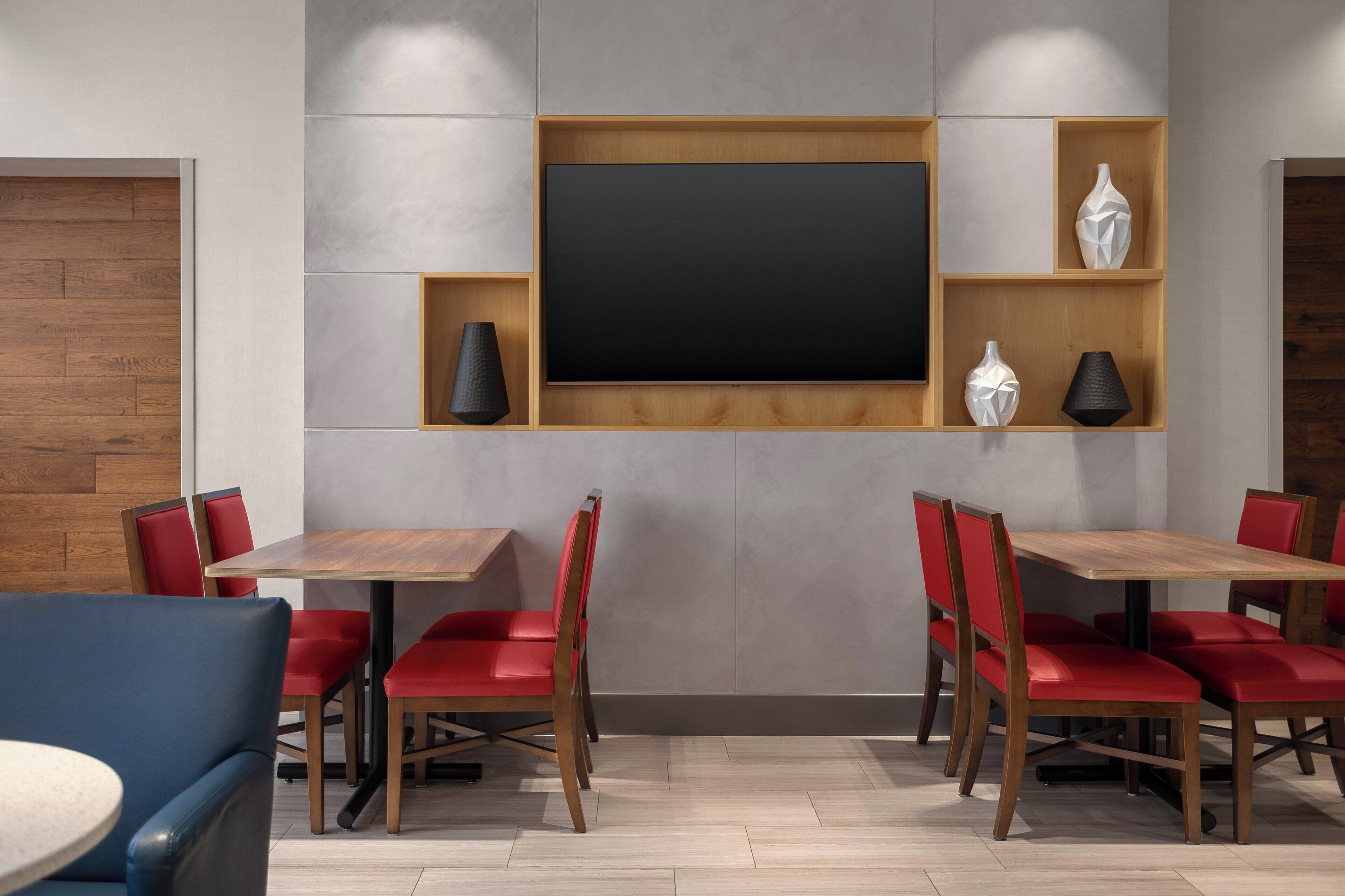 A modern dining area with a large flat-screen TV, wooden tables, red chairs, and decorative vases.