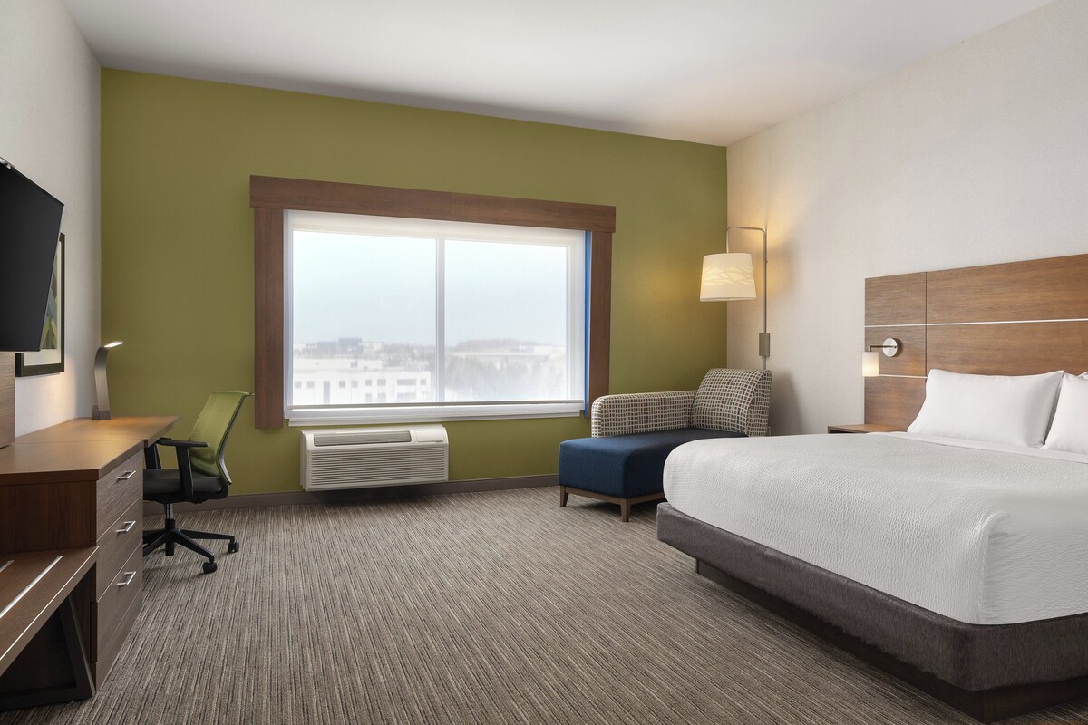 A hotel room with a bed, a desk, a chair, a window with a view, and a wall-mounted lamp.