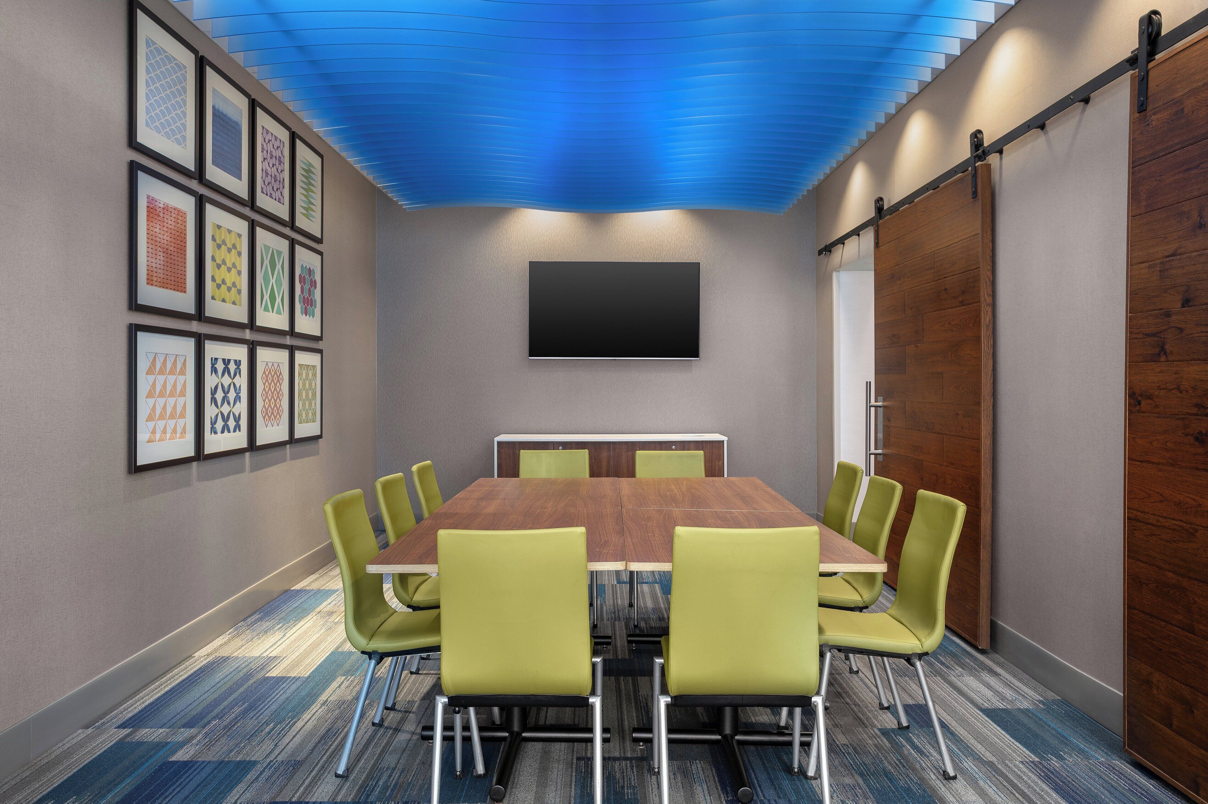 A conference room with a large table, green chairs, a mounted TV, and framed artwork on the wall.