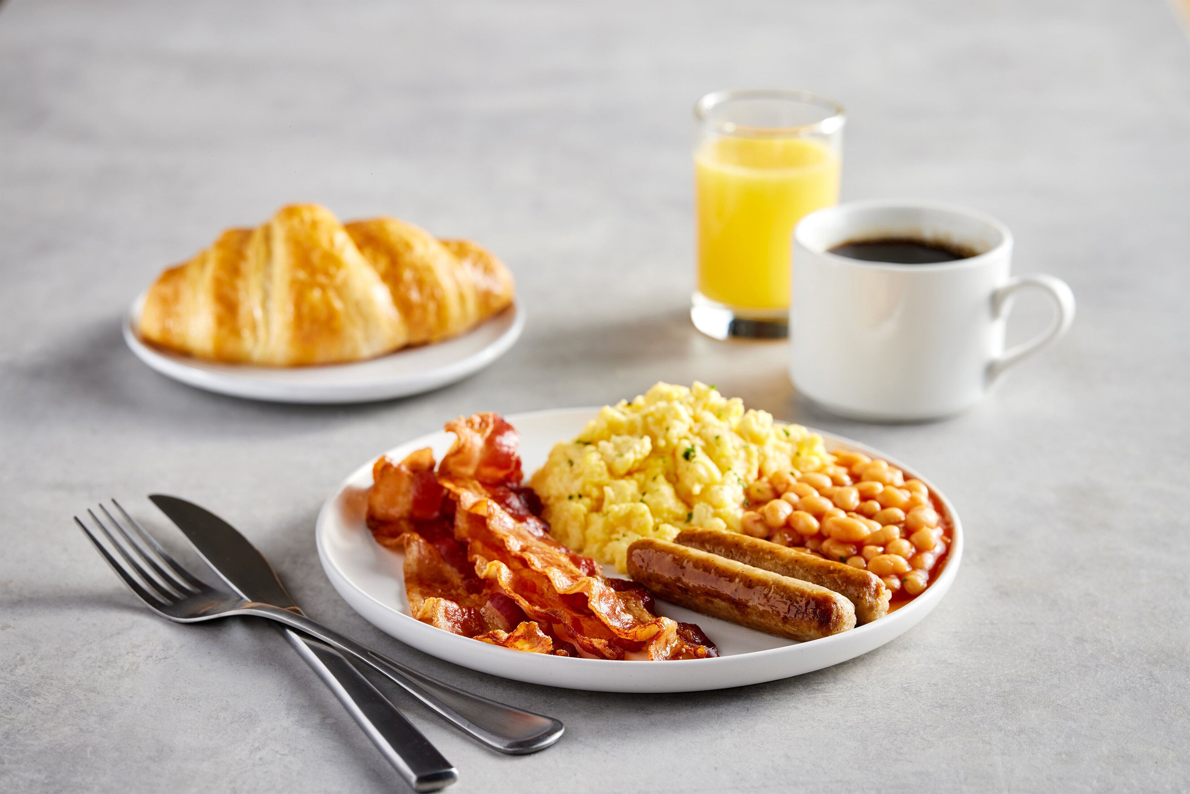 A breakfast plate with eggs, bacon, sausages, beans, and a croissant on a white plate, accompanied by a glass of orange juice and a cup of coffee.