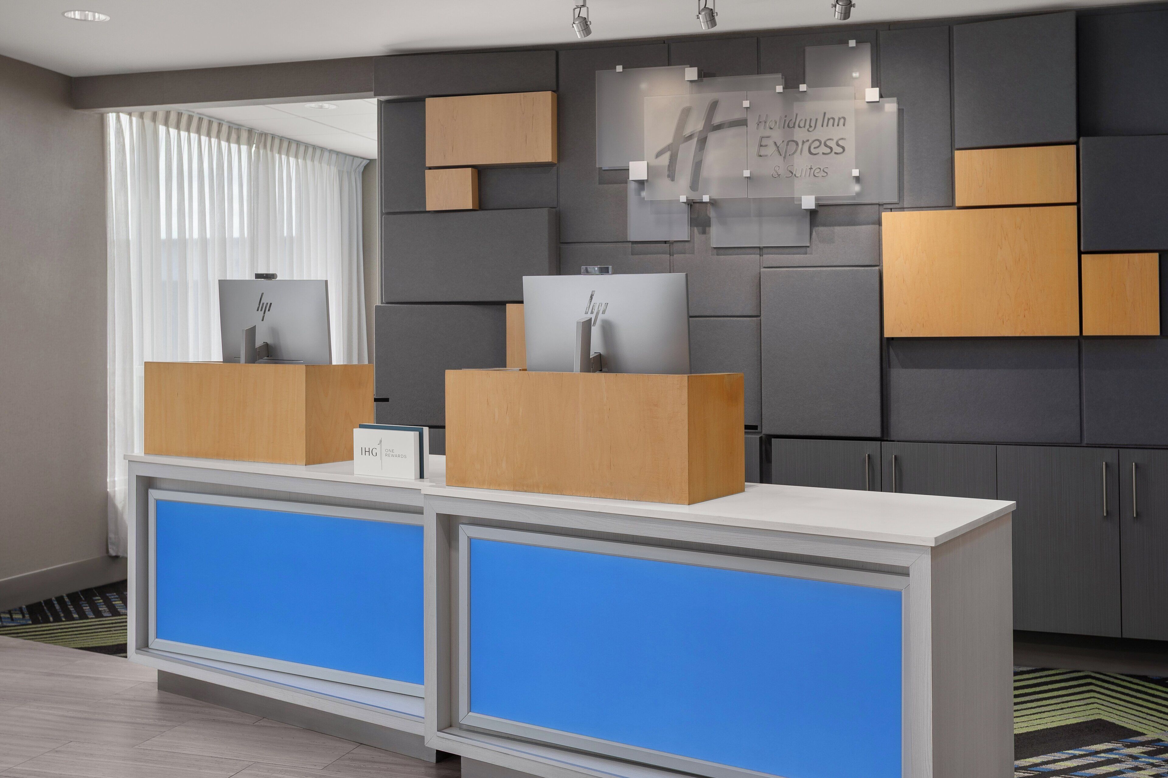 A reception desk with a blue panel, a sign for Holiday Inn Express Suites, and a window with vertical blinds.
