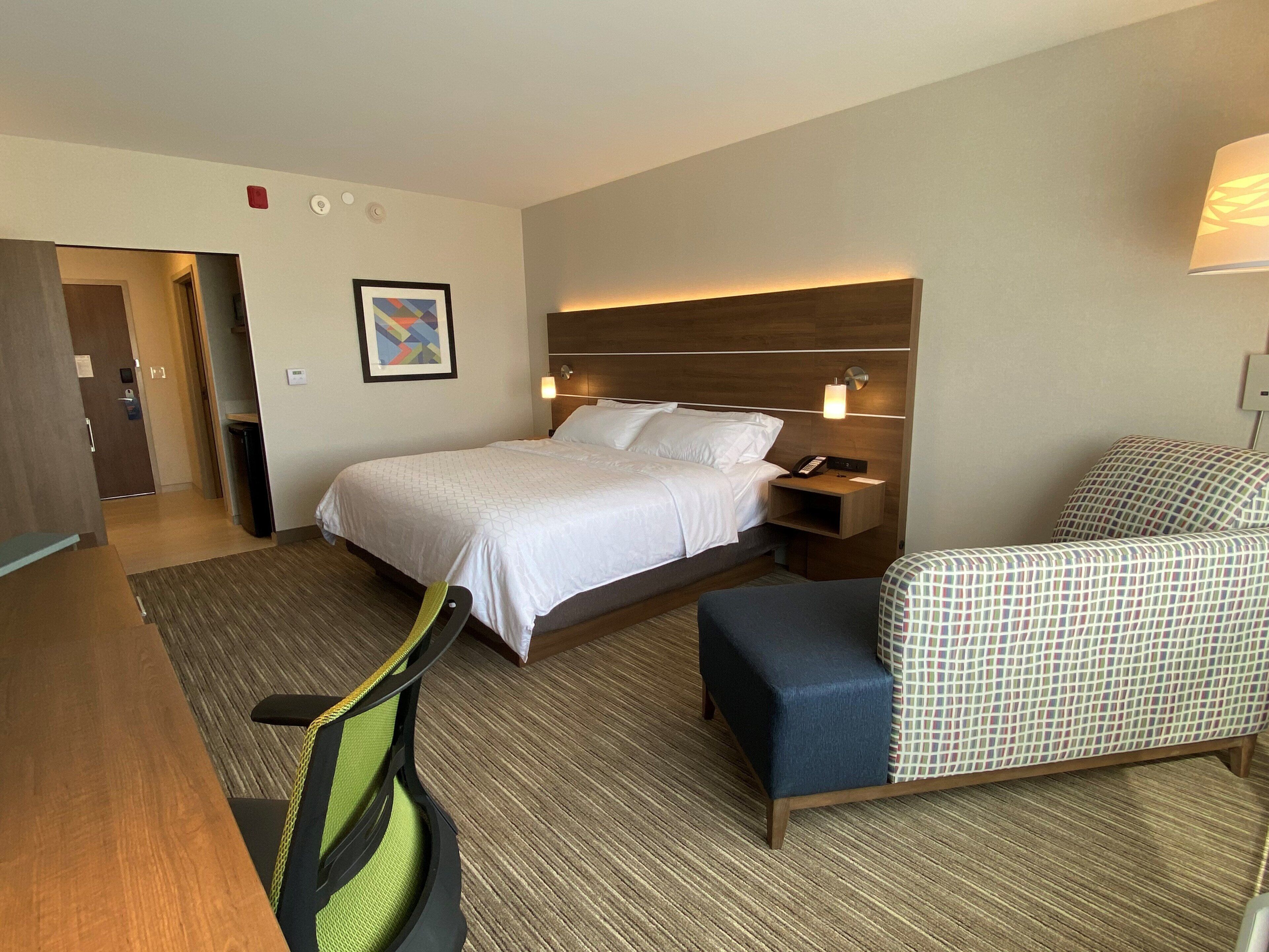 A hotel room with a bed, bedside table, chair, desk, and a framed picture on the wall.