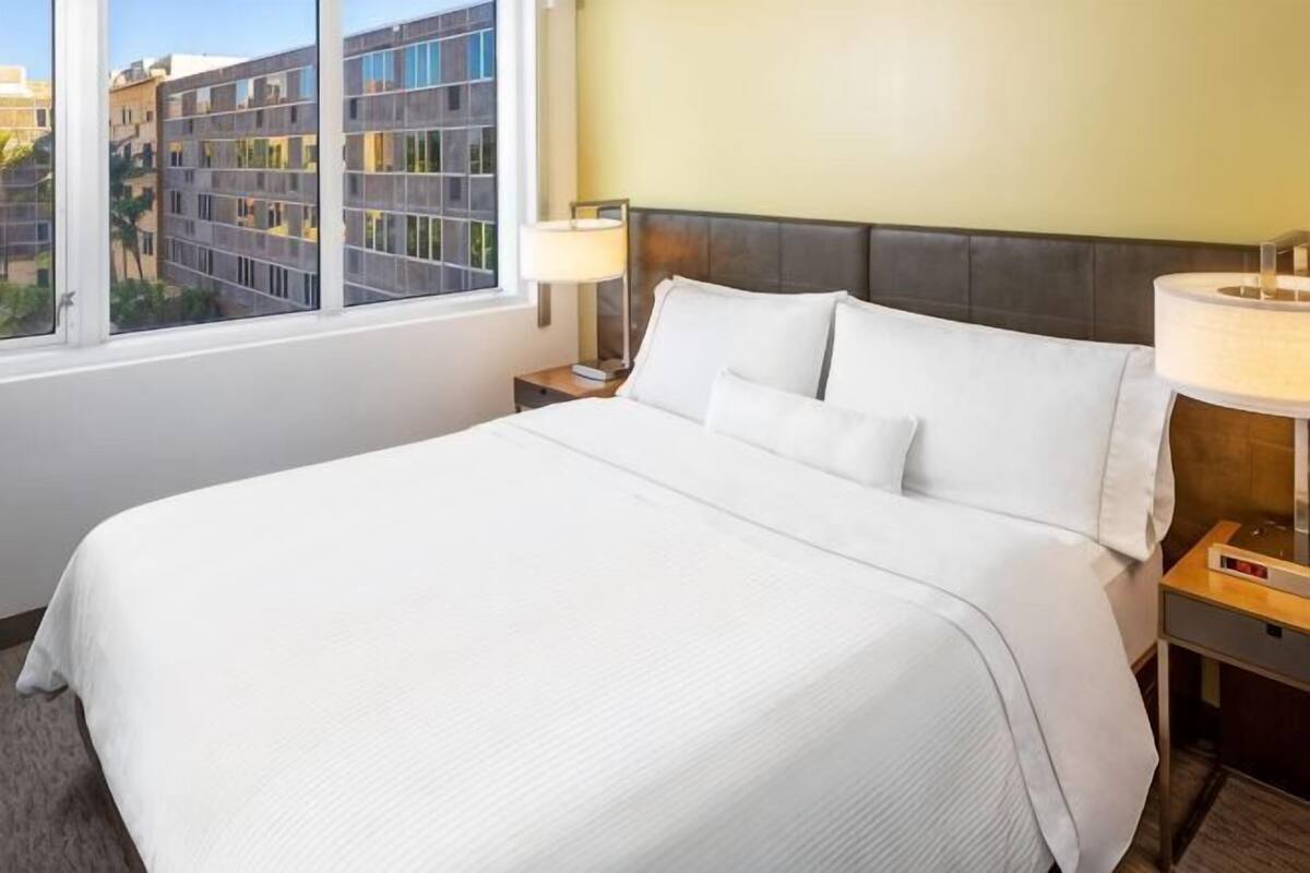 A hotel room with a large bed, a nightstand, a lamp, and a view of buildings through a window.