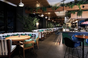 A restaurant with wooden tables and chairs, green cushioned seating, hanging plants, and a bar area. - Virgin Hotels Nashville (Nashville)