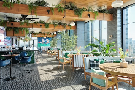 A modern outdoor seating area with wooden tables and chairs, surrounded by large windows and potted plants.