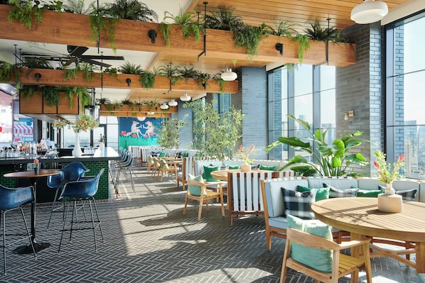 Un espacio moderno al aire libre con mesas y sillas de madera, rodeado de grandes ventanales y plantas en macetas.