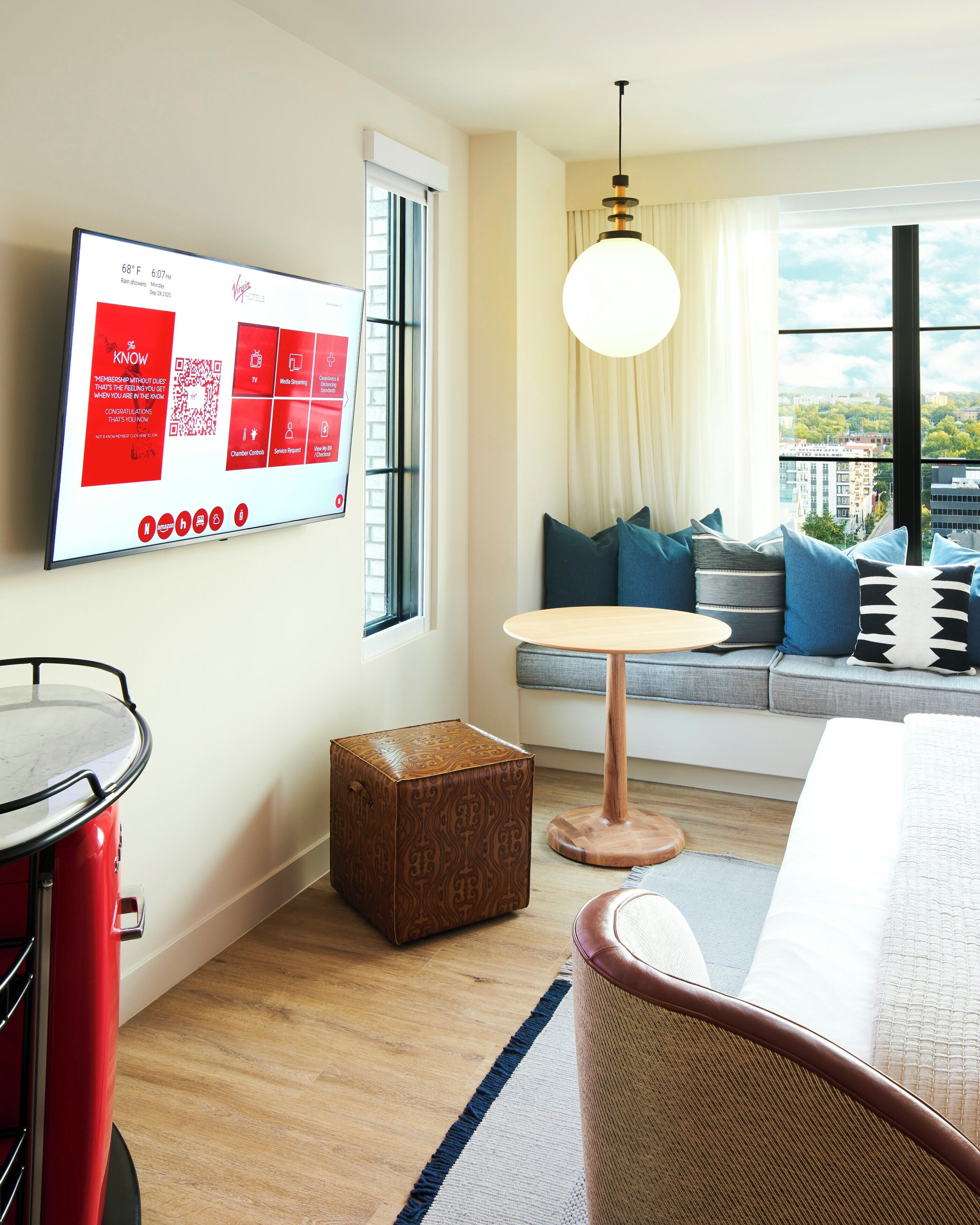 A modern hotel room with a large TV displaying a welcome message, a cozy seating area with cushions, a round table, and a view of the city through large windows.