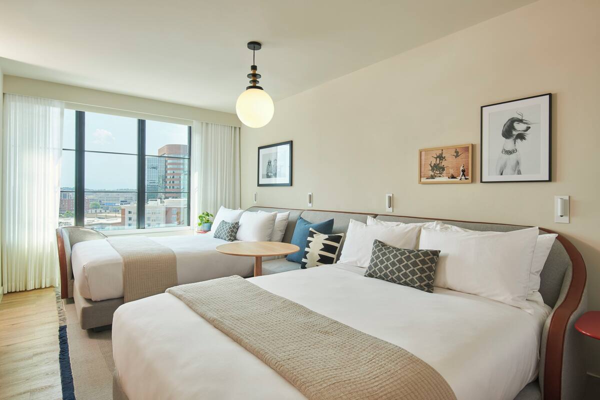 Ein Hotelzimmer mit zwei Betten, einem runden Tisch und großen Fenstern mit Blick auf die Stadt.