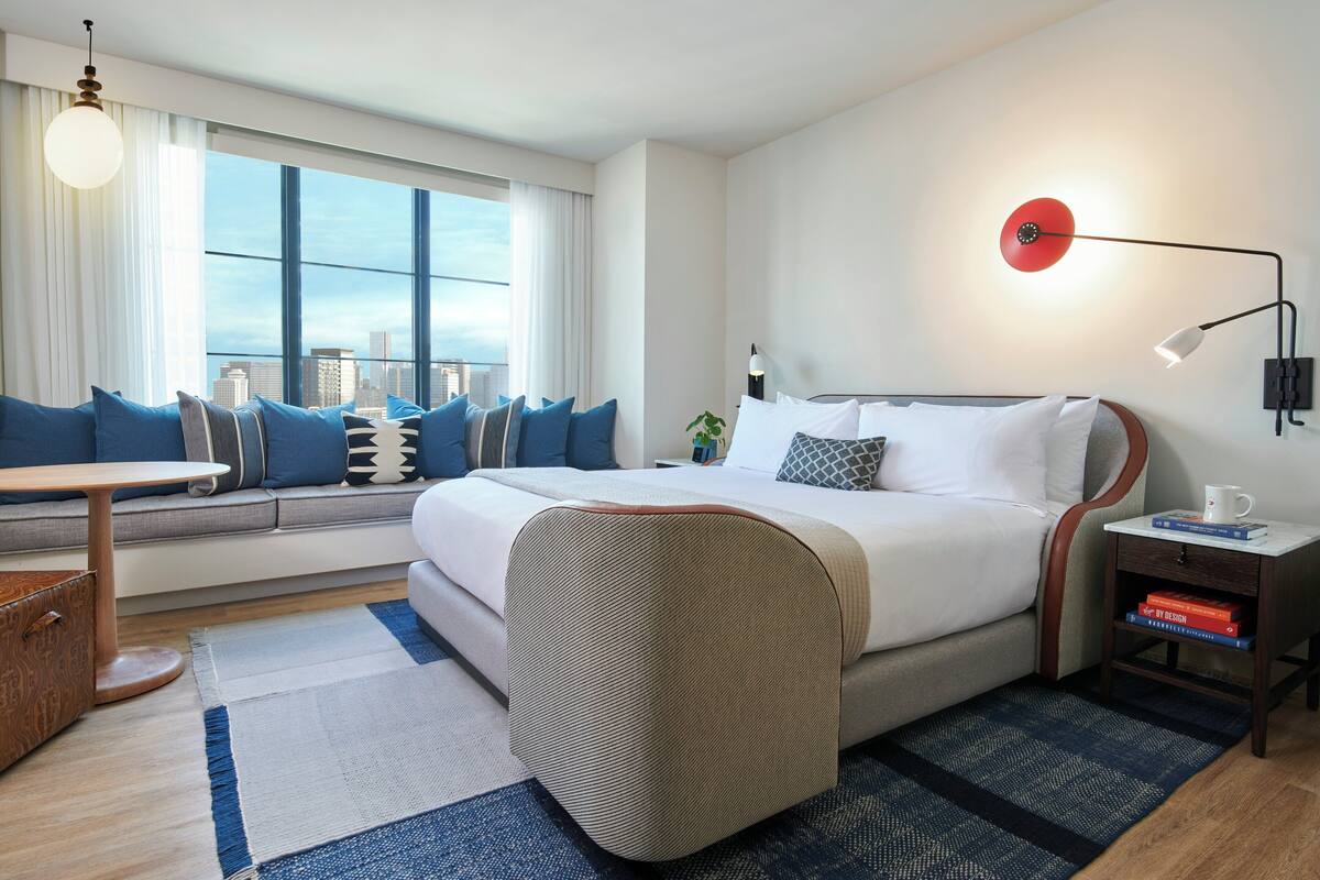 Ein modernes Hotelzimmer mit einem großen Bett, einer Sitzecke mit Sofa und kleinem Tisch, einem Blick auf die Stadt durch große Fenster und einer roten Wandleuchte.