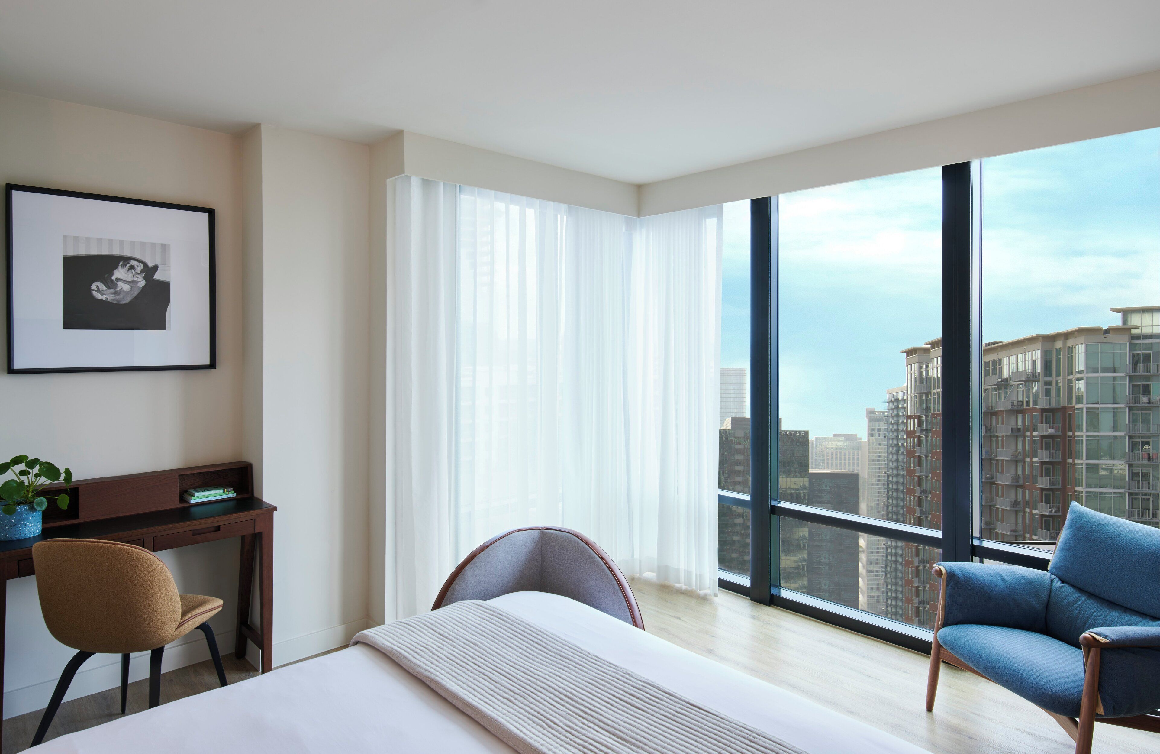 Een moderne hotelkamer met een groot bed, een bureau, een stoel en een bank. Er is een groot raam met uitzicht op hoge gebouwen.