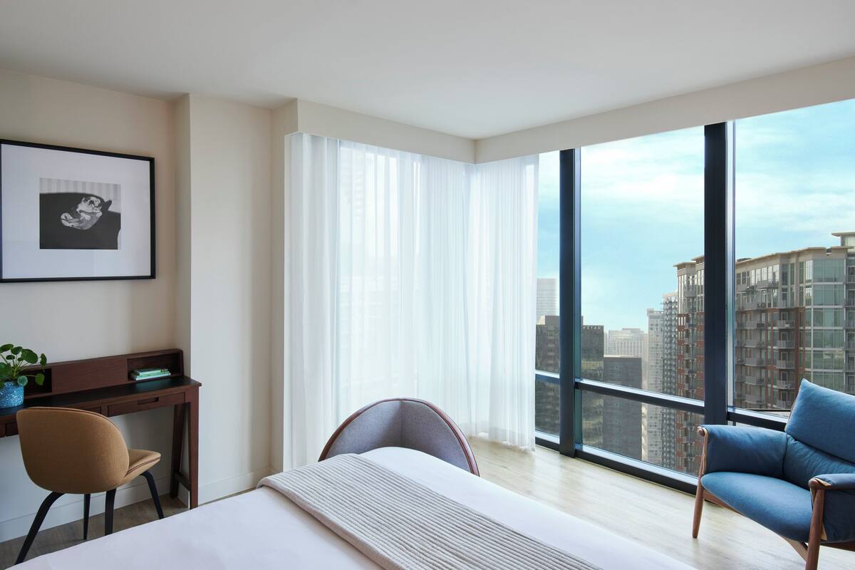 Ein modernes Hotelzimmer mit einem großen Bett, einem Schreibtisch, einem Sessel und einer Couch. Ein großes Fenster bietet Blick auf hohe Gebäude.