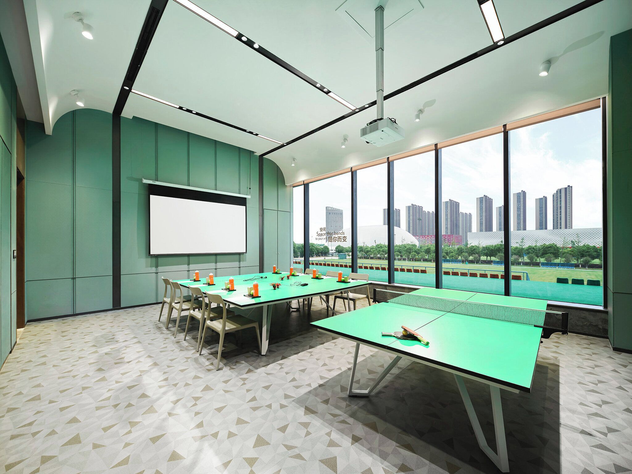Una sala de conferencias con una gran mesa, sillas, una pantalla de proyector y vistas a un paisaje urbano a través de amplias ventanas.