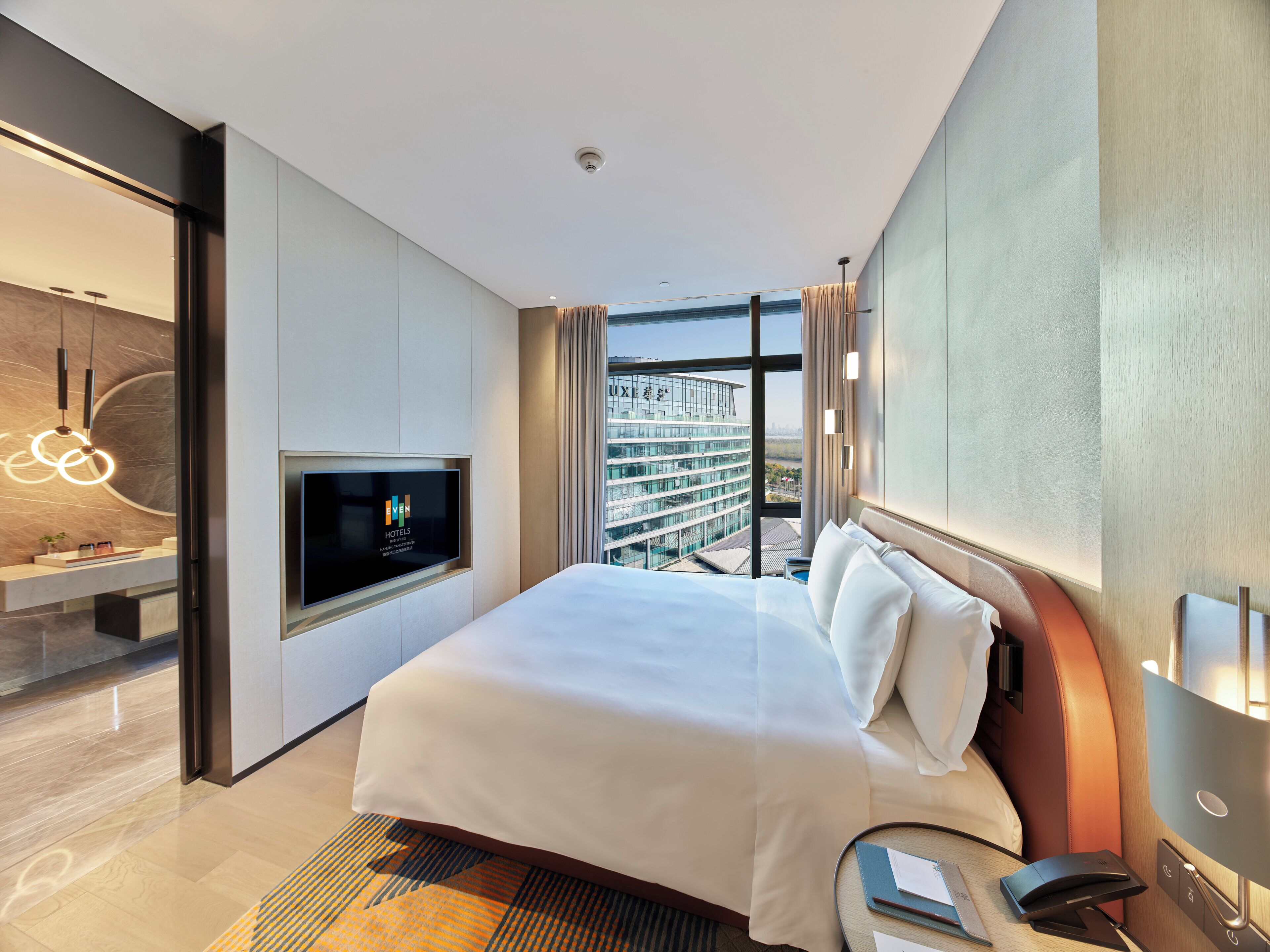 Ein modernes Hotelzimmer mit einem großen Bett, einem Fernseher, einem Schreibtisch mit Lampe und Blick auf die Stadt.