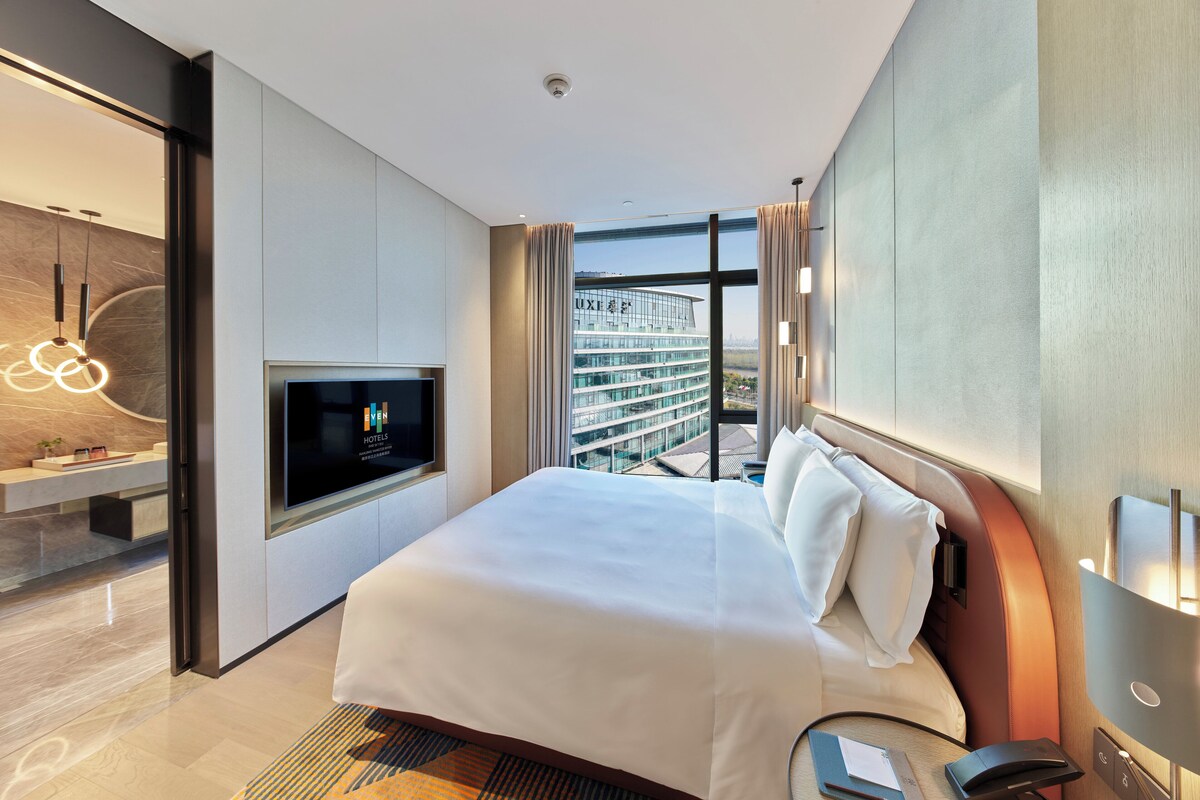 A modern hotel room with a large bed, a TV, a desk with a lamp, and a view of a cityscape.