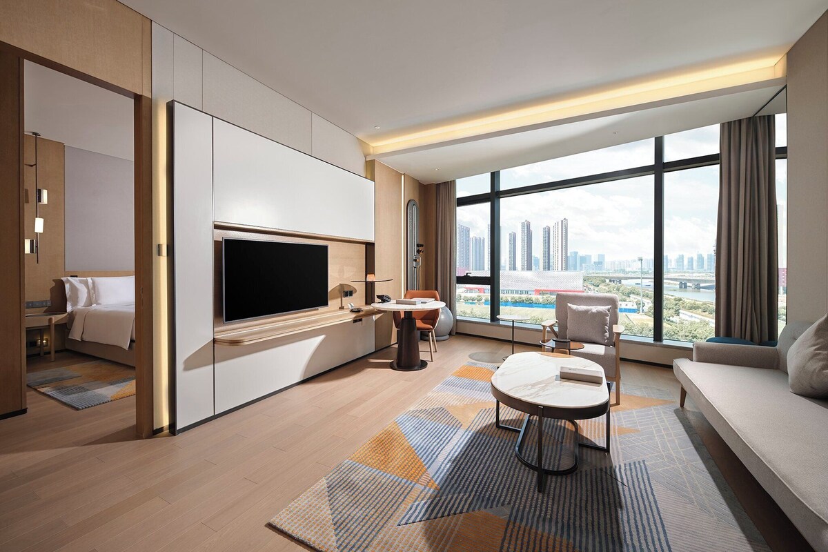 A modern hotel room with a large TV, a sofa, a dining table, and a view of the city skyline.