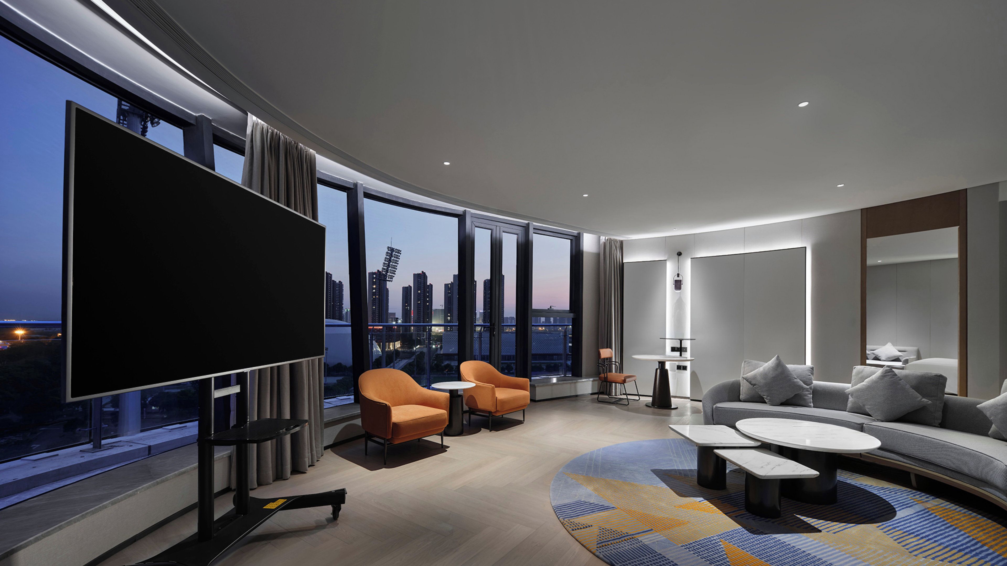 Habitación de hotel moderna con una televisión grande, asientos cómodos, una pequeña mesa y vistas a la ciudad.