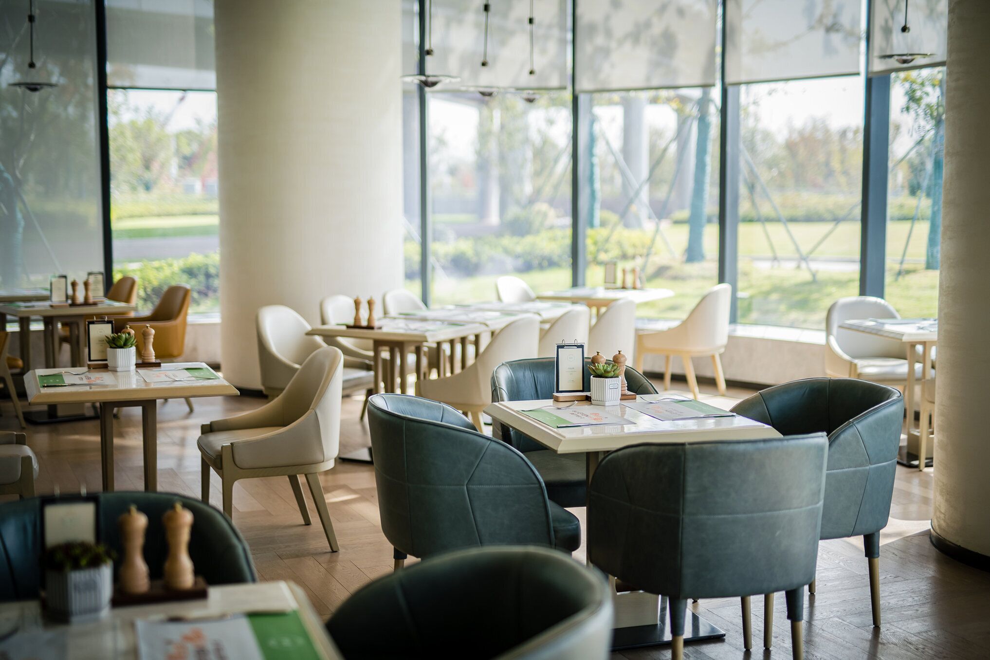 A modern dining area with tables and chairs, large windows, and a view of greenery outside.