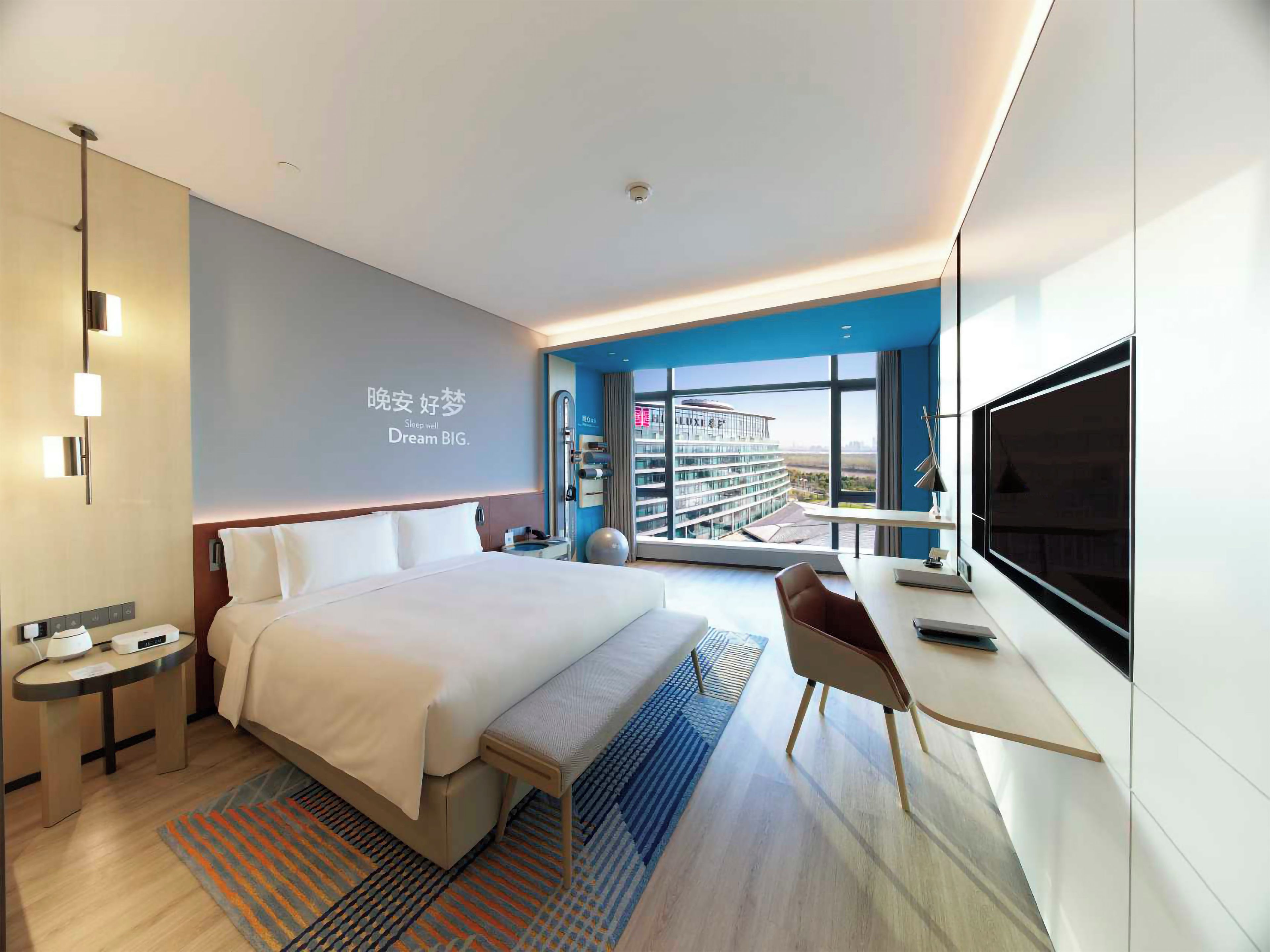 A modern hotel room with a large bed, a desk, a television, and a view of a building.