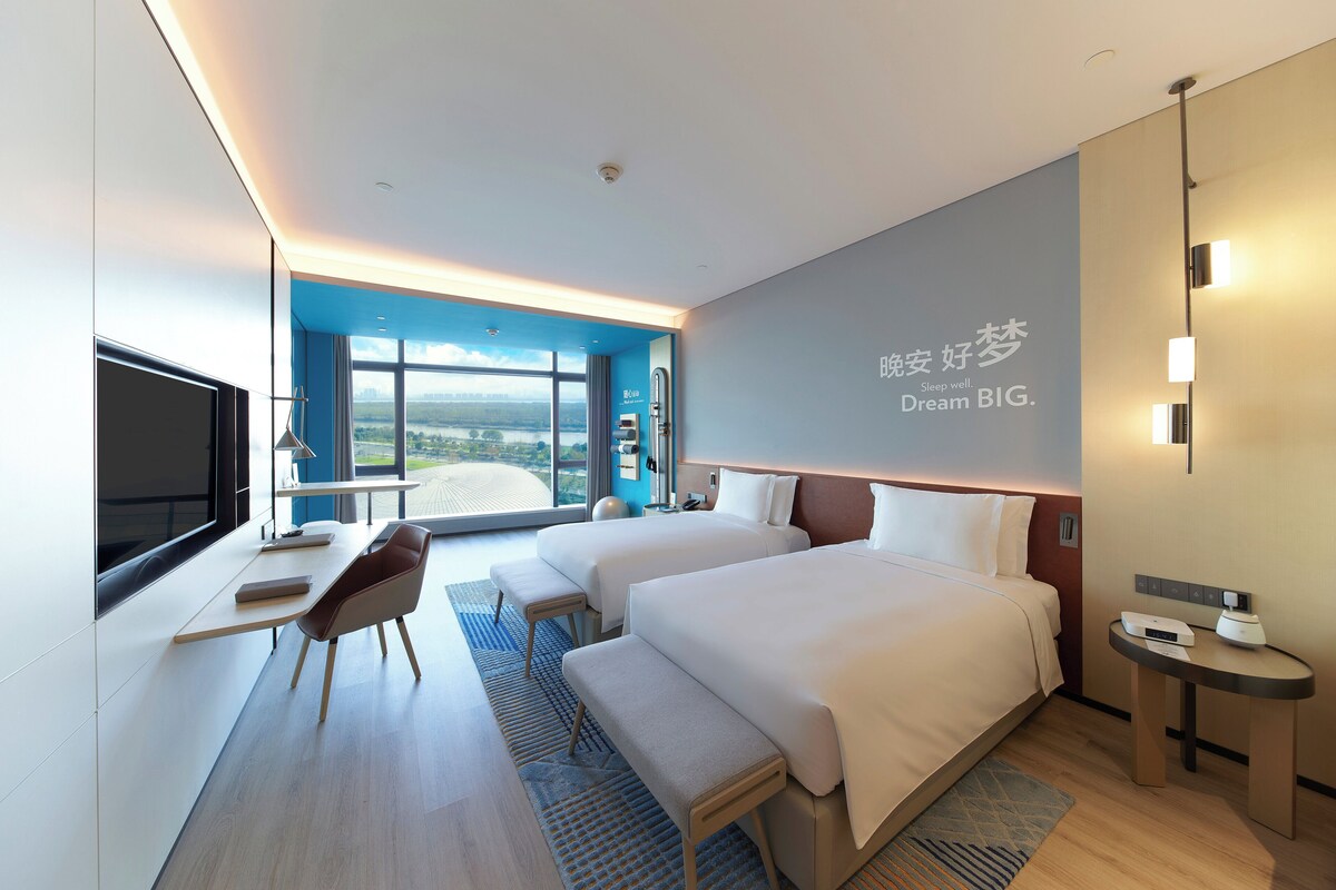 A modern hotel room with two beds, a desk, a chair, a TV, and a large window offering a city view.