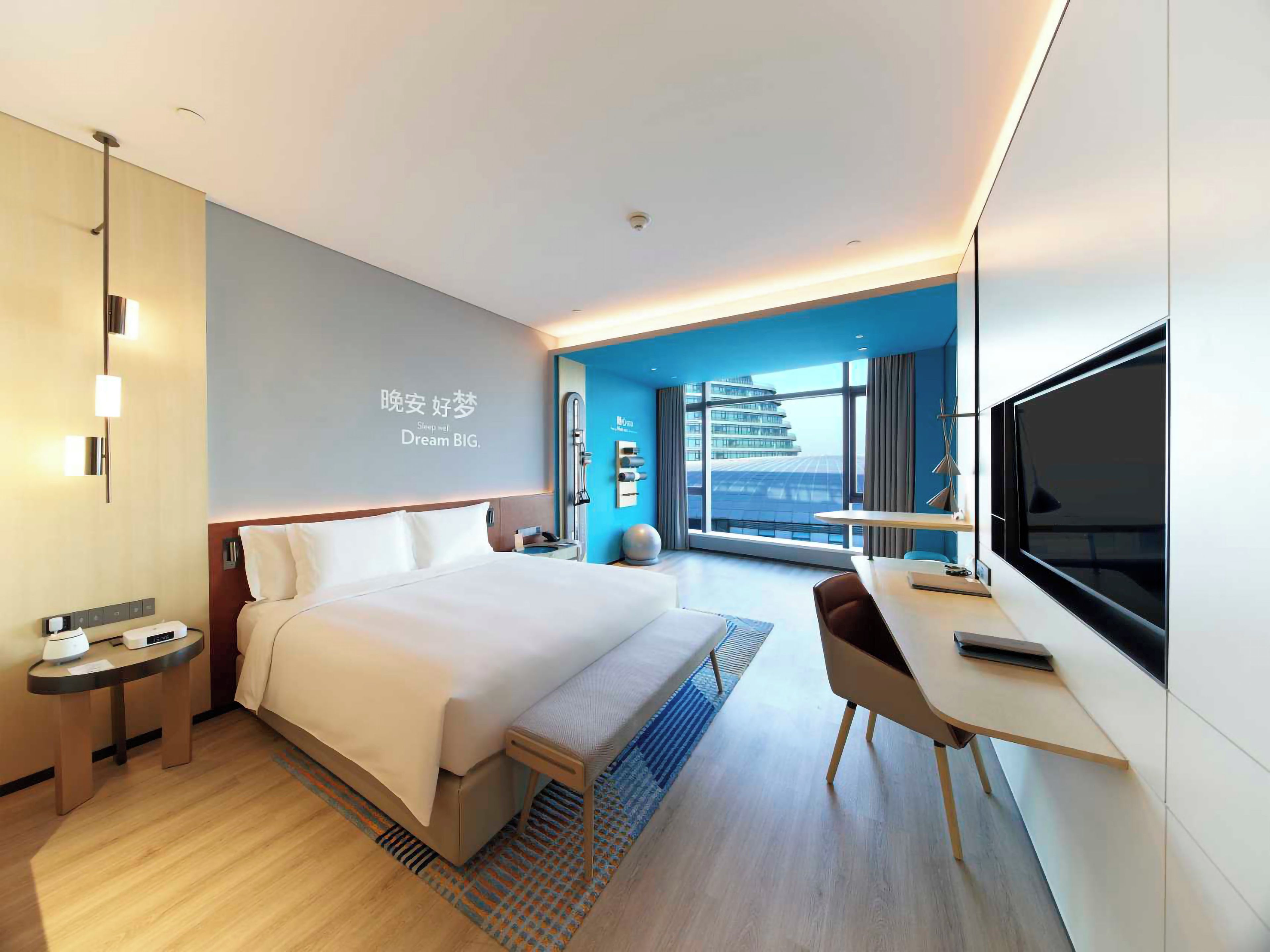 Una habitación de hotel moderna con cama, mesita de noche, escritorio, silla y un amplio ventanal con vista a la ciudad.