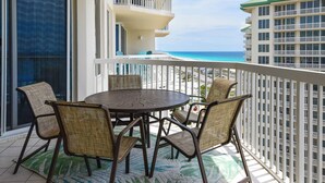 Een balkon met een ronde tafel en vier rieten stoelen, met uitzicht op een strand en een hotelgebouw.