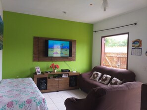 Una sala de estar con una pared verde, un televisor montado en la pared, un mueble de madera con altavoces, un sofá y una cama con ropa de cama estampada.
