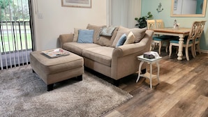 Een woonkamer met een beige bank, een kleine tafel en een eethoek met een houten tafel en stoelen.