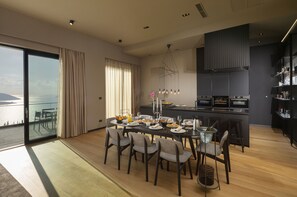 Un'elegante zona pranzo con un tavolo grande apparecchiato per sei persone, una cucina con mobili scuri e vista mare attraverso ampie finestre.