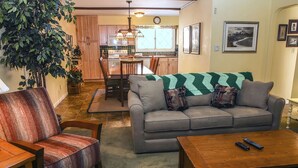 A living room with a grey sofa, a wooden coffee table, and a striped armchair. There is a dining table with chairs and a green blanket. A large plant is in the corner, and a clock is on the wall. - Fawn Valley Inn: 240 (Estes Park)