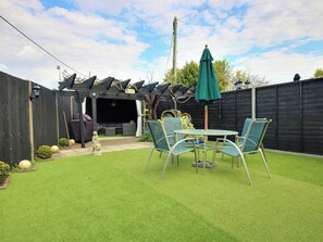 Een achtertuin met kunstgras, een patio tafel met stoelen, een groene parasol en een houten schutting.