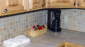 A kitchen with wooden cabinets, a tiled backsplash, a coffee maker, a basket with snacks, and a folded towel on the counter. - Fawn Valley Inn: 292 (Estes Park)