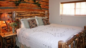 A cozy bedroom with a rustic log wall, a bed with patterned pillows, a bedside table with a lamp and vase, and a window with blinds. - Fawn Valley Inn: 300 (Estes Park)