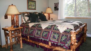 A rustic wooden bed with a quilt, two bedside lamps, and a window with a view of trees. - Fawn Valley Inn: 306 (Estes Park)
